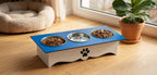 A raised pet feeder and water bowl stand placed on a wooden floor in a bright room. The stand is white with a blue top surface, three bowls (two filled with dry food and one empty), a black paw print detail on the front, and is styled near a window with a potted plant and a pet bed in the background.