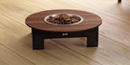 A round wooden pet fire table with a dark brown wood-grain top, a small drawer, and a stainless steel fire bowl filled with rocks, set on a light wood plank floor.