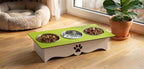 A raised pet feeder and water fountain stand with a bright green top, white base, and three bowls (two food bowls, one water bowl). It is shown on a wooden floor in a home setting next to a potted plant and a pet bed.