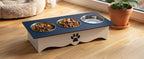 A raised pet feeder and waterer stand with a dark blue top, white base, and a black paw print logo. It holds two bowls of dry food and one empty water bowl, set on a light wood floor next to a potted plant.