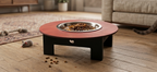A raised pet feeder table with a round reddish-brown wooden top, black legs, and a stainless steel bowl filled with kibble, set on a wooden floor in a living room with a cat nearby.