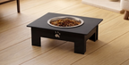 A black elevated pet feeder stand with a stainless steel bowl filled with dry dog food, placed on a light wood floor. The stand features a small white paw print detail on the front.