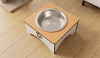 A square elevated pet feeder stand with a light wood top, white base, and a stainless steel bowl, shown on a light wood plank floor.